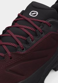 Gros plan sur une chaussure de randonnée rouge foncé et noire avec une languette rembourrée, des lacets rouges et noirs à motifs, et un petit logo blanc sur la languette.