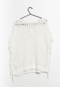 Poncho en tricot blanc avec un tissage ouvert, des bords à franges et une forme rectangulaire ample. Suspendu à un cintre noir.