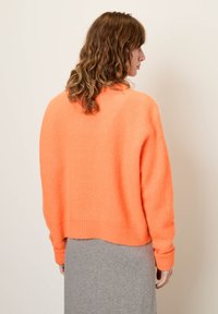 Prevelik, oranž pulover iz mehkega, teksturiranega materiala z rebrastimi manšetami in robom; stiliziran z sivim krilom za kontrast.