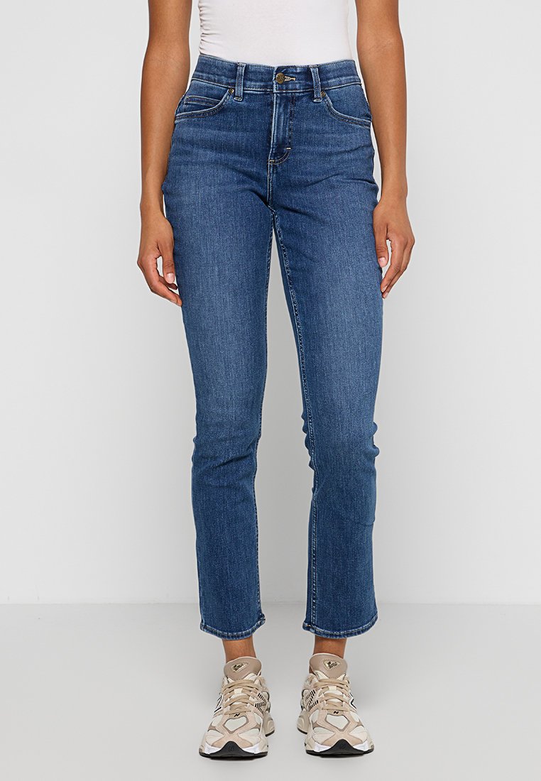 Jeans w kolorze niebieskim o wysokim stanie, dopasowanej konstrukcji i zwężanych nogawkach, z lekkim przetarciem na kostkach. Noszone z beżowymi trampkami.
