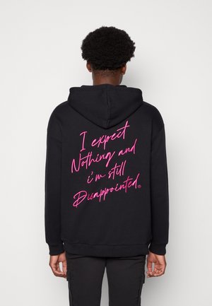 Zwarte hoodie met roze cursieve tekst op de achterkant die leest: "Ik verwacht niets en ben nog steeds teleurgesteld," met een ruime capuchon en boorden.
