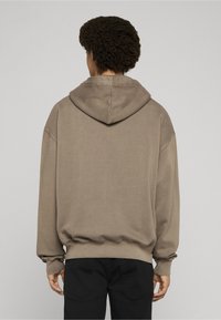 Person med krøllete hår iført en løs brun hettegenser og svarte bukser, stående med ryggen vendt mot en ensfarget lys bakgrunn.