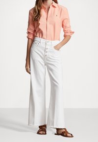 Světle růžová košile na knoflíky, bílé džínové kalhoty s vysokým pasem a širokým střihem s třepeným lemem, hnědé sandály na řemínky. Klasický casual outfit.