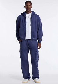 Navy blue hoodie with large front pockets, paired with matching cargo pants featuring buttoned side pockets and white shoes.