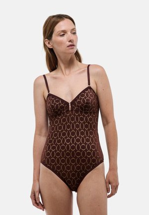 Femme portant un maillot de bain une pièce marron avec un motif ovale beige et de fines bretelles, regardant vers sa gauche sur un fond uni.