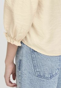 Krämfärgad texturerad blus med rynkade ärmar, matchad med ljusblå denimjeans som har en bakficka och en blekt finish.