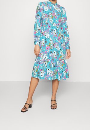 Frau trägt ein blaues Midikleid mit Blumenmuster, langen Ärmeln und gerüschtem Saum, kombiniert mit schwarzen Sandalen mit Absatz, steht vor einem neutralen Hintergrund.