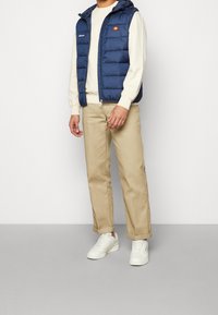 Námořnicky modrá prošívaná vesta s kapucí, s červenými a žlutými logy na hrudi, v kombinaci s krémovou mikinou a béžovými kalhotami a bílými teniskami.