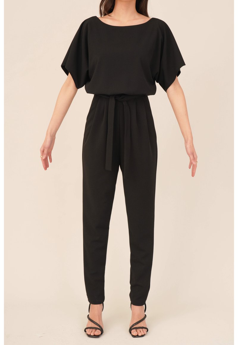 Femme portant une combinaison noire à manches courtes avec une ceinture à la taille, un pantalon fuselé et des sandales à talons hauts noires à lanières, debout les bras légèrement écartés.