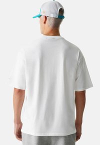 Camiseta blanca de algodón con mangas cortas, vista trasera lisa, combinada con pantalones de chándal grises. También es visible una gorra de malla blanca con acentos turquesa.