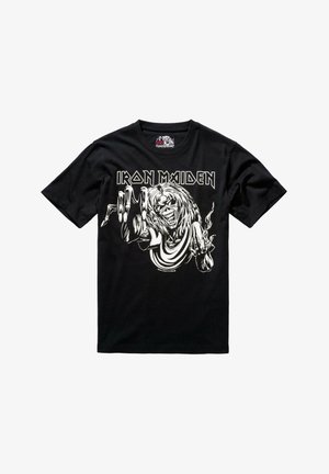Melna kokvilnas T-kreklā ar lielu baltu grafiku, kurā attēlots skeleta figūra ar gariem matiem, kas pacēlusi roku, virs kura ir teksts "IRON MAIDEN".