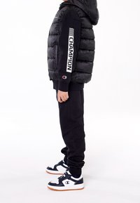 Czarna pikowana kamizelka z kapturem, czarna bluza z białym napisem "CHAMPION" na rękawie, czarne spodnie dresowe oraz granatowo-białe trampki z logo.