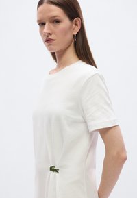 Λευκό βαμβακερό t-shirt με κοντά μανίκια, που φέρει ένα πράσινο λογότυπο κροκόδειλου στο πλαϊνό μέρος, με λεία υφή και άνετη εφαρμογή.