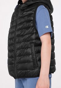 Schwarze, gepolsterte Weste mit Kapuze, mit gestepptem Design und Reißverschluss. Getragen über einem blauen Hemd, das auf beiden Teilen ein Logo zeigt.