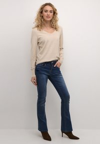 Pull beige à col en V associé à un jean bootcut bleu. Le modèle porte des escarpins bruns à bout pointu. Le pull a des poignets côtelés ; le jean a des poches classiques.