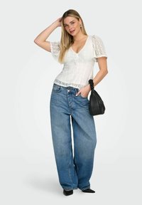 Jeune femme portant un haut en dentelle blanche, un jean bleu ample, des chaussures noires à bout pointu, tenant un sac à main noir avec une poignée froncée, posant avec la main sur la tête.