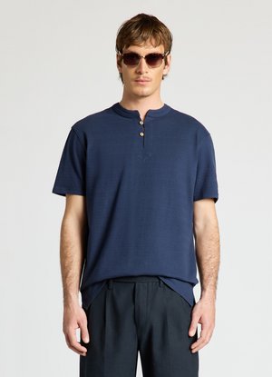 Mann mit dunkler Sonnenbrille, marineblauem kurzärmeligem Henley-Shirt mit zwei Knöpfen und dunkler Hose, stehend vor schlichtem hellem Hintergrund.