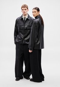 Melnas mākslīgās ādas jakas ar priekšējām kabatām, vilktas pār brīvām melnām biksēm. Modeļa detaļas ietver strukturētas apkakles un gludu tekstūru.