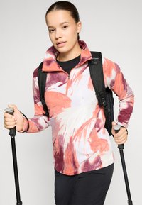 Fleece pullover met een kwart ritssluiting, voorzien van een multikleurig abstract patroon in tinten oranje en bordeauxrood, gecombineerd met een zwarte rugzak.