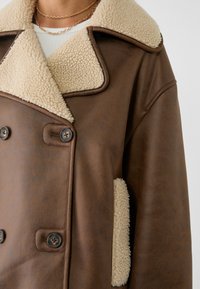 Veste en shearling double boutonnage marron avec doublure en polaire crème au col et aux poches, portée sur un haut blanc avec un collier en chaîne dorée.