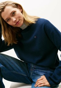 Junge Frau mit blonden Haaren, die einen marineblauen Pullover über einem grauen Rollkragenpullover und blauen Jeans trägt, sitzt entspannt vor einem weißen Hintergrund.