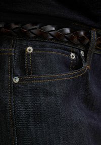 Nærbillede af en mørkeblå denimjeanslomme med orange syninger, metalnitter og et flettet mørkebrunt læderbælte.