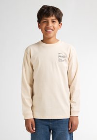 Beige langärmliges T-Shirt aus Baumwolle mit einem kleinen gedruckten Logo auf der linken Brust, kombiniert mit blauen Denim-Jeans.