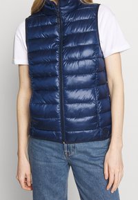 Marineblauer Steppweste mit hohem Kragen, gestepptem Design, Frontreißverschluss und glatter Textur. Trägt man über einem weißen T-Shirt und Jeans.