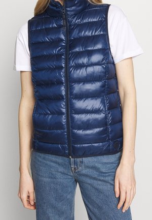 Gilet rembourré bleu marine avec un col haut, un design matelassé, une fermeture éclair sur le devant et une texture lisse. Porté sur un t-shirt blanc et un jean en denim.