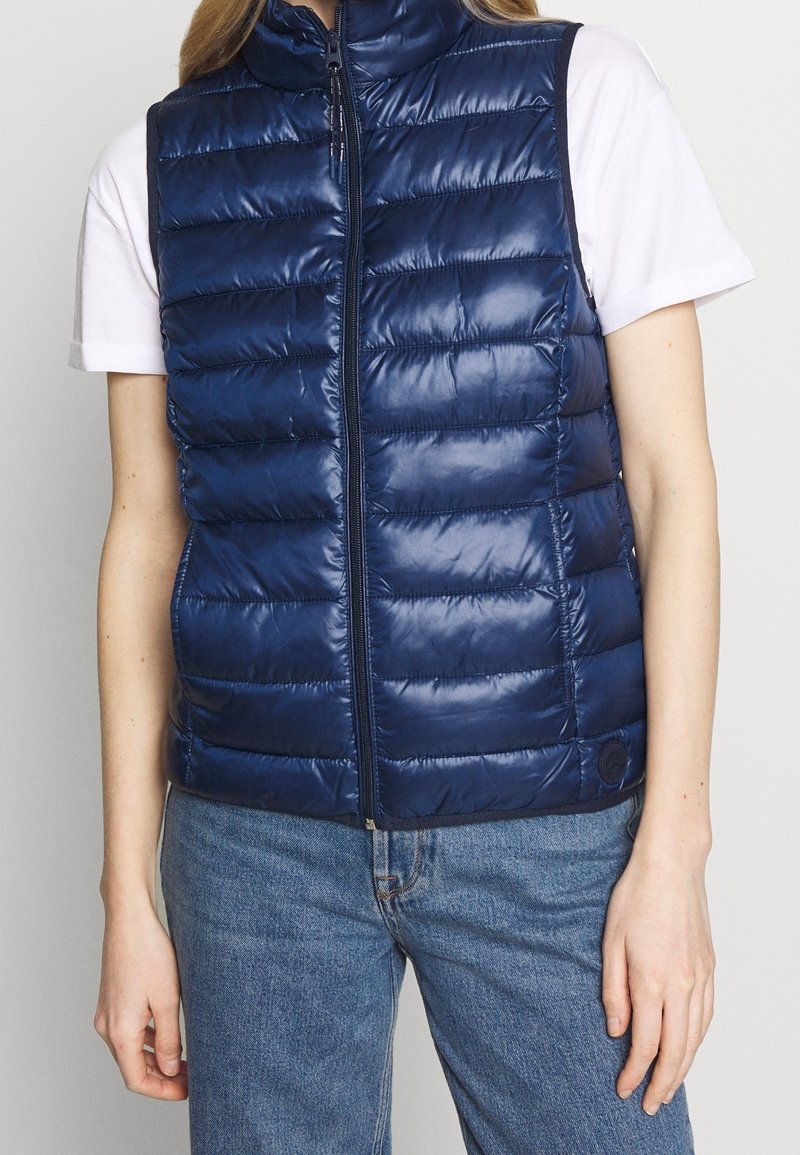 Marineblauer Steppweste mit hohem Kragen, gestepptem Design, Frontreißverschluss und glatter Textur. Trägt man über einem weißen T-Shirt und Jeans.