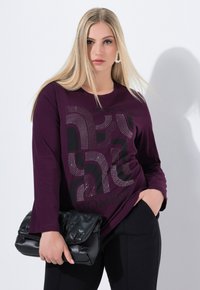 Musta bluuvi, jossa on pitkät hihat ja violetti väri, mustalla printtimusterilla, jossa on kaarevia viivoja ja jalokivitehosteita, yhdistettynä mustaan, pullotettuun clutch-laukkuun.