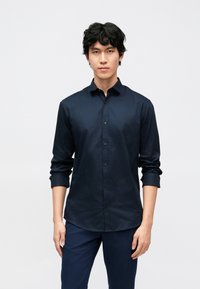 Jonge man met zwart krullend haar, gekleed in een marineblauw overhemd met knopen en marineblauwe broek, staand tegen een effen lichte achtergrond.