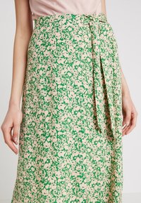 Blommig omlottskjol med ett grönt och beigt mönster, tillverkad av lätt tyg med en knytning vid midjan.