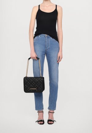 Frau trägt ein schwarzes Tanktop, blaue High-Waist-Jeans, schwarze Absatzsandalen und hält eine schwarze gesteppte Love Moschino Handtasche mit Kettenriemen.