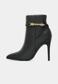 Botín negro de tacón alto con punta afilada y detalle de correa con hebilla dorada alrededor de la parte superior.