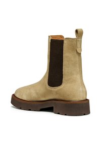 Bota de tobillo de ante beige con un panel lateral elástico marrón, punta redondeada y suela de goma texturizada para mayor agarre. Asas en la parte trasera.