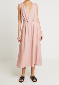 Rosa, ärmlös A-linjeformad klänning med djup V-ringning och midjedetaljer som har två knappar. Tyget har en strukturerad yta.