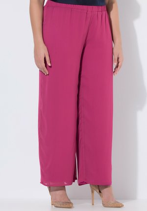 Frau trägt locker sitzende, hoch taillierte magentafarbene Hosen mit elastischem Bund und goldenen High Heels, steht vor einem weißen Hintergrund.