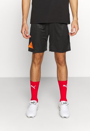 Pantalones cortos deportivos negros con un acabado texturizado, con un parche del logo de Puma en color naranja. Combinados con calcetas rojas hasta la rodilla y botas blancas de fútbol.