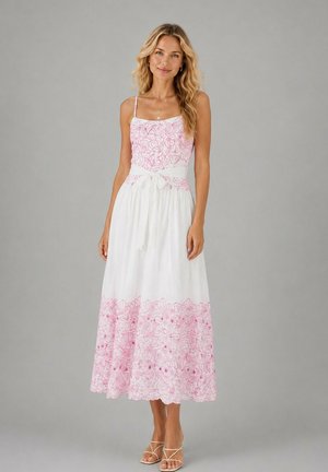 Femme aux longs cheveux blonds portant une robe blanche sans manches avec une broderie florale rose et une ceinture à la taille, debout devant un fond gris.