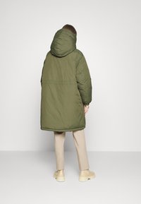 Grön puffjacka med huva, med en lös passform och sömmande detaljer. Modellen har på sig beige byxor och skor, sedd bakifrån.