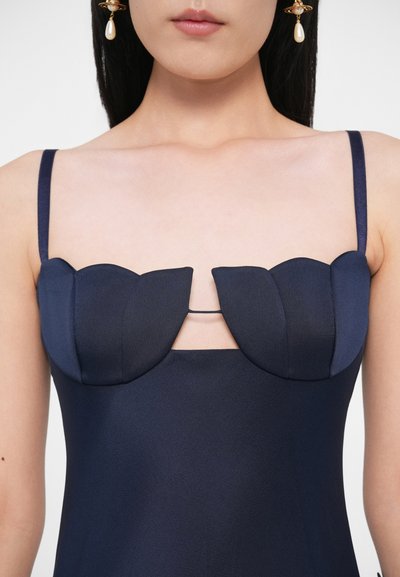 Sieviete tērpusies tumši zilā kleitā ar strukturētām viļņotām krūzītēm un plānām plecu lencītēm, valkā zelta auskarus ar pērļu piliņām.