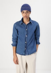 Camicia in denim blu con bottoni, colletto e maniche arrotolate, con bottoni in argento e una finitura morbida e testurizzata. Pantaloni beige indossati sotto.