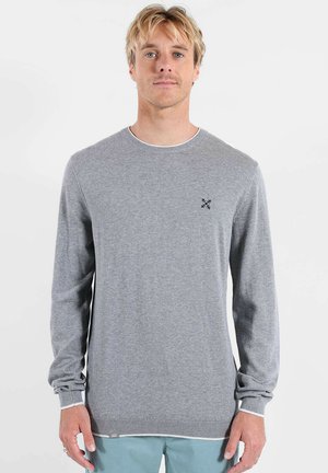 Homme aux cheveux blonds portant un pull gris clair à col rond avec un petit logo brodé sur la poitrine gauche, debout devant un fond uni.