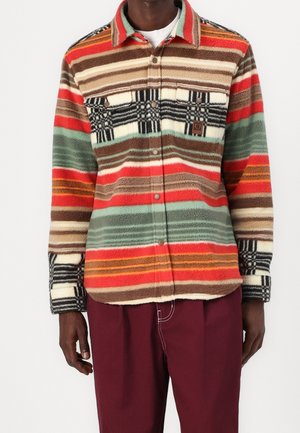 Homme portant une veste polaire rayée multicolore avec des poches boutonnées et un pantalon bordeaux sur un fond uni.