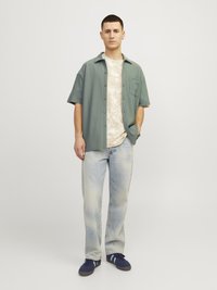 Kortærmet, lys grøn button-up skjorte over en beige mønstrede t-shirt, parret med lyseblå jeans og marineblå sneakers.