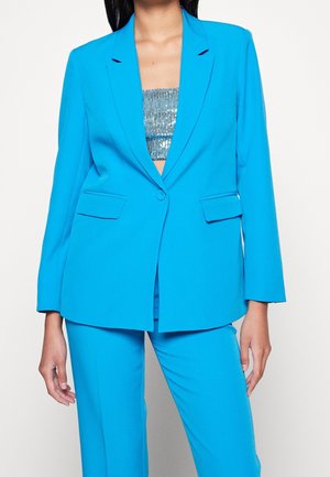 Femme portant un blazer bleu vif sur mesure et un pantalon assorti, avec un haut bandeau argenté à sequins en dessous, se tenant devant un fond uni.