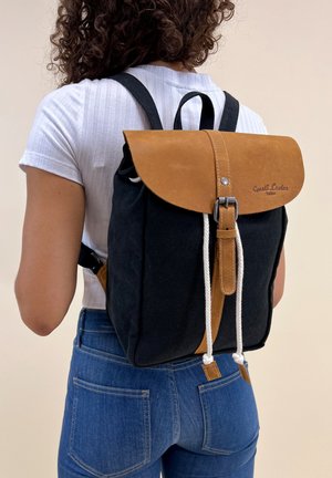 Zaino in tela con patta in pelle marrone, chiusura con cordoncino bianco e dettaglio del marchio. Corpo nero con accenti beige, spallacci regolabili.