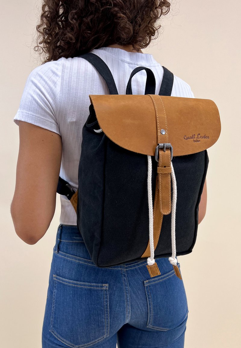Zaino in tela con patta in pelle marrone, chiusura con cordoncino bianco e dettaglio del marchio. Corpo nero con accenti beige, spallacci regolabili.