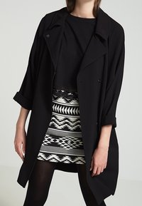 Cappotto oversize nero con ampi rever, bottoni doppi e maniche arrotolate, indossato sopra una gonna a motivo bianco e nero.